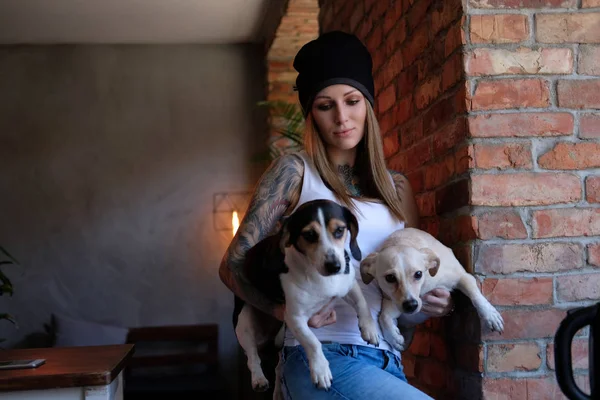 Retrato de una chica tatuada hipster vestida con camiseta blanca y sombrero sostiene a sus perritos en la habitación con el interior del loft . —  Fotos de Stock