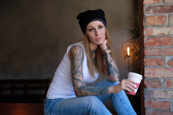 Dalgın tattoed hippi kız beyaz gömlek giymiş ve çatı iç ile bir odada masada otururken paket servisi olan restoran kahve şapka tutar. — Stok fotoğraf