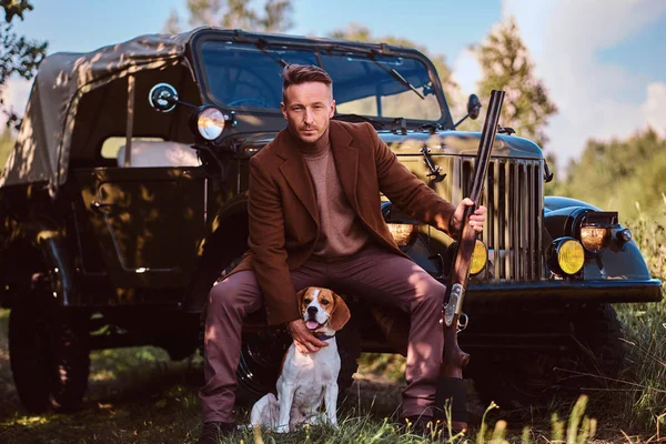 Jägaren i eleganta kläder håller ett hagelgevär och sitter tillsammans med sin beaglehund medan lutande mot en retro militära bil i skogen. — Stockfoto