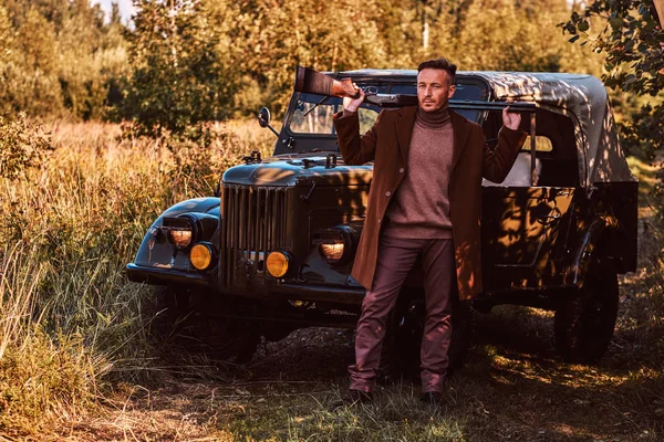 Jäger in eleganter Kleidung hält eine Schrotflinte in der Hand und steht mit einem Retro-Militärauto im Wald. — Stockfoto