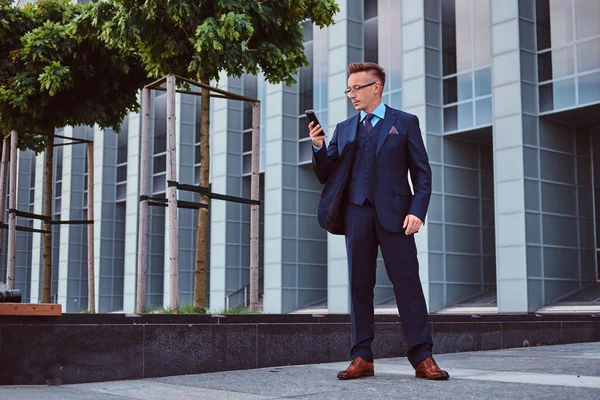 Retrato de un hombre de negocios con estilo y confianza vestido con un traje elegante usando un teléfono inteligente mientras está parado al aire libre contra el fondo del rascacielos . —  Fotos de Stock