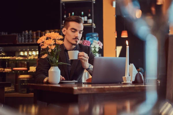 Hombre freelancer guapo con barba elegante y cabello vestido con un traje negro que trabaja en el ordenador portátil mientras está sentado en un café . —  Fotos de Stock