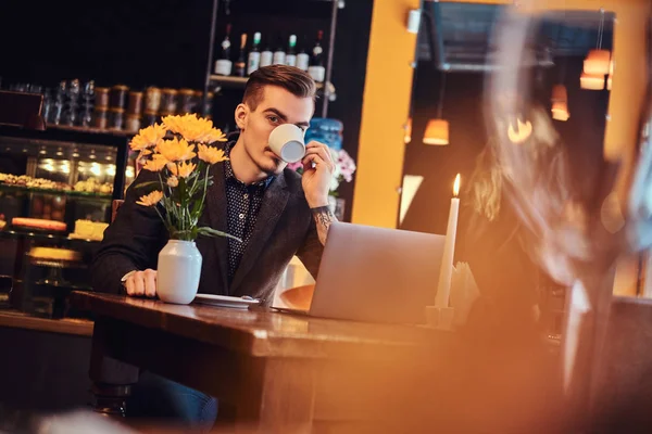 Freelancer όμορφος άνθρωπος με στιλάτα γένια και μαλλιά ντυμένος με ένα μαύρο κοστούμι που κάθεται σε ένα καφέ με ένα ανοιχτό laptop και πίνει καφέ, κοιτάζοντας την κάμερα. — Φωτογραφία Αρχείου