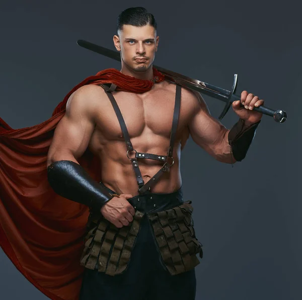 古代ギリシャの戦士の戦闘服に身を包んだ筋肉質ボディの肖像画は彼の肩に剣を保持します 灰色の背景に分離 — ストック写真