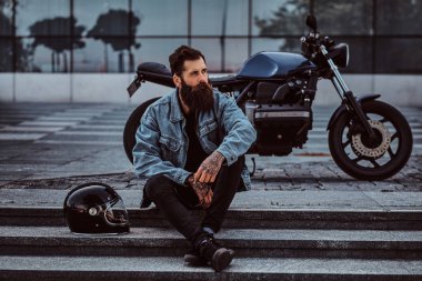 Acımasız bir sakallı erkek portresi adımlar yakınındaki bir gökdelen karşı özel yapım retro motosiklet üzerinde oturan bir kot ceket giymiş.