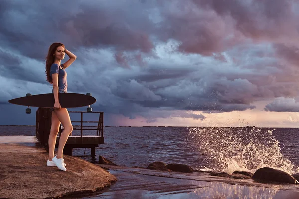 Retrato de una chica sensual sostiene un monopatín mientras está de pie en la playa está disfrutando de un clima nublado oscuro increíble durante el atardecer . — Foto de Stock