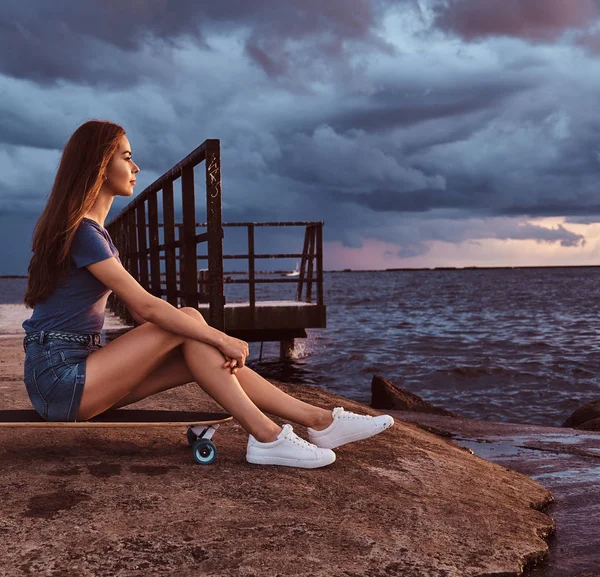 Menina sensual sentado em um skate na praia está desfrutando de incrível clima nublado escuro durante o pôr do sol . — Fotografia de Stock