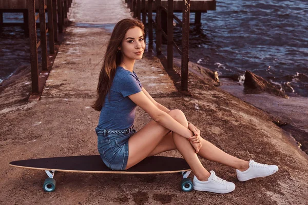 Portrait of a girl sitting on a skateboard on the old pier is enjoying amazing dark cloudy weather during sunset. — Stock Photo, Image