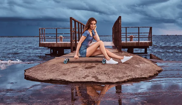 Menina sensual sentado em um skate na praia está desfrutando de incrível clima nublado escuro durante o pôr do sol . — Fotografia de Stock