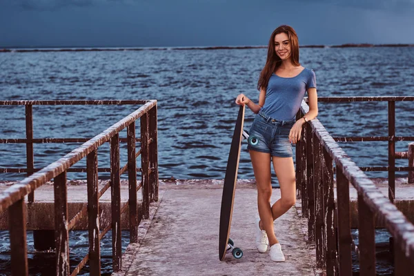 Menina sensual posando com skate em pé no cais velho está desfrutando de incrível clima nublado escuro durante o pôr do sol . — Fotografia de Stock