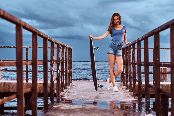 Sensuale ragazza in posa con skateboard in piedi sul vecchio molo sta godendo di incredibile buio nuvoloso tempo durante il tramonto . — Foto Stock