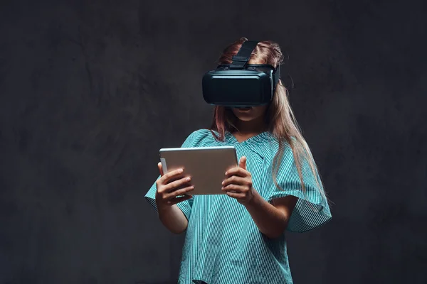 Jong meisje met behulp van virtuele realiteit brillen en bezit van een tablet-pc. Geïsoleerd op donkere gestructureerde achtergrond. — Stockfoto