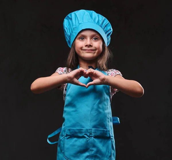 Feliz niña vestida con uniforme de cocinero azul muestra gesto de amor. Aislado sobre un fondo de textura oscura . —  Fotos de Stock