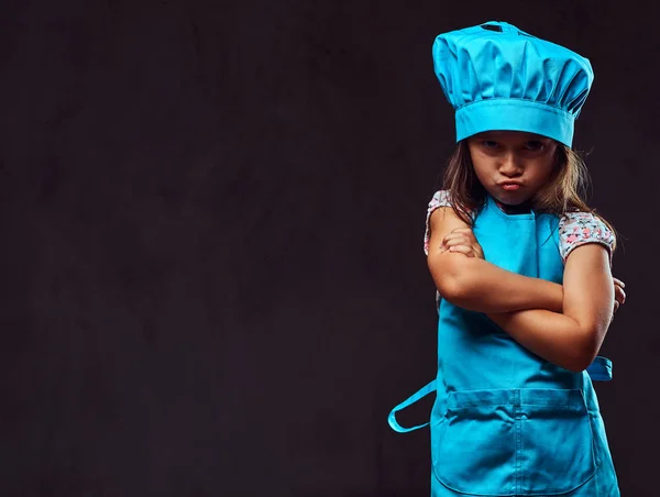 Niña descontenta vestida con uniforme de cocinero azul de pie con los brazos cruzados. Aislado sobre fondo de textura oscura . —  Fotos de Stock