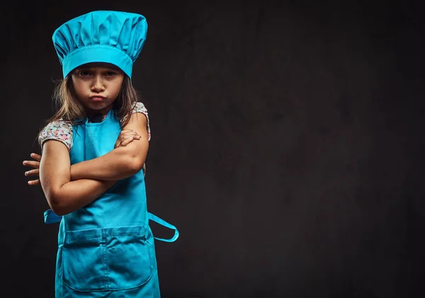 Hoşnutsuz küçük kız mavi aşçı Tekdüzen ayakta çapraz silah ile giyinmiş. Karanlık dokulu arka plan üzerinde izole. — Stok fotoğraf