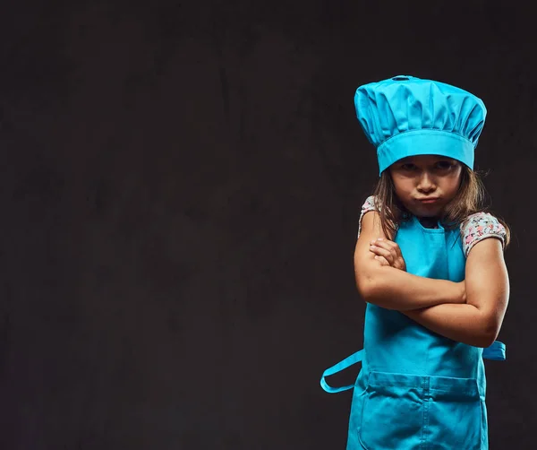 Niña descontenta vestida con uniforme de cocinero azul de pie con los brazos cruzados. Aislado sobre fondo de textura oscura . —  Fotos de Stock