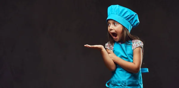 Sorprendida niña vestida de cocinero azul posando en un estudio. Aislado sobre fondo de textura oscura . —  Fotos de Stock