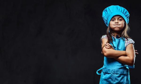 Bambina scontenta vestita in uniforme blu cuoco in piedi con le braccia incrociate. Isolato su sfondo scuro testurizzato . — Foto Stock
