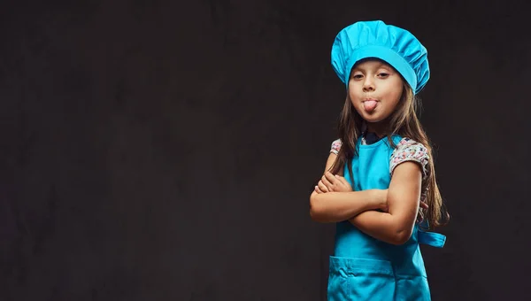 Недовольная маленькая девочка, одетая в синюю форму повара, стоит со скрещенными руками и показывает вырванный язык. Изолированный на темном текстурированном фоне . — стоковое фото