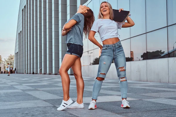 Zwei lustige Hipster-Mädchen, die mit einem Skateboard auf dem Hintergrund des Wolkenkratzers stehen. — Stockfoto