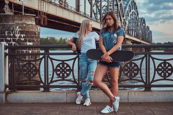 Zwei schöne Hipster-Mädchen, die mit Skateboard gegen eine Brücke stehen. — Stockfoto