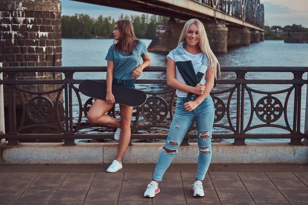 Zwei schöne Hipster-Mädchen, die mit Skateboard gegen eine Brücke stehen. — Stockfoto