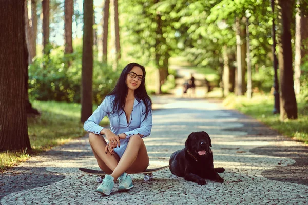 戴眼镜的快乐性感黑发女孩穿着衬衫和短裤坐在滑板上 与她的狗在公园散步 — 图库照片