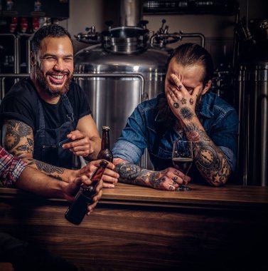 İki sakallı Interracial arkadaş, bir bira fabrikasında zanaat bira içmek. Apronda dövmeli hipster çalışan erkek arkadaşı bir bira fabrikası ile bira içiyor.