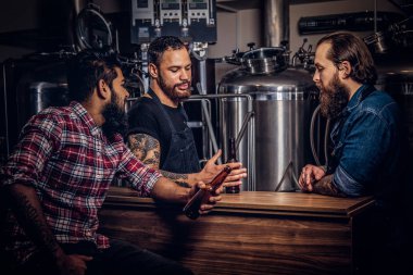 Üç sakallı Interracial arkadaş, bir bira fabrikasında zanaat bira içmek. Önlük iki hipster işçi ile onların arkadaşı bir bira fabrikasında bira içmek.