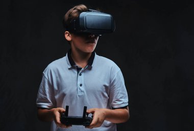 Genç bir çocuk portresi sanal gerçeklik gözlükle beyaz bir t-shirt giymiş ve denetim uzak tutar. Koyu arka plan üzerinde izole.