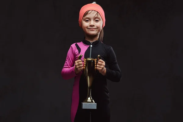 Happy little skater girl champion holds a winners cup. Isolated on dark textured background.