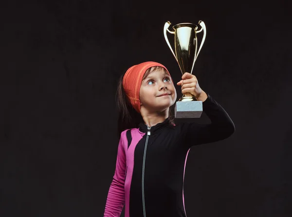 Happy little skater girl champion holds a winners cup. Isolated on a dark textured background.