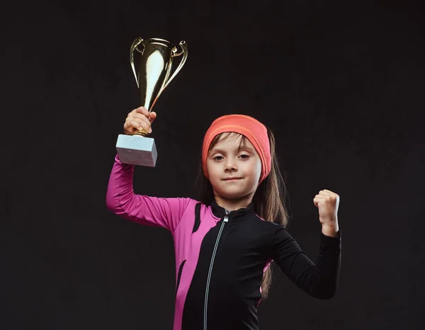 Happy little skater girl champion holds a winners cup. Isolated on a dark textured background.