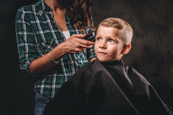 Children hairdresser with the trimmer is cutting little boy against a dark background. Cute preschooler boy getting haircut.