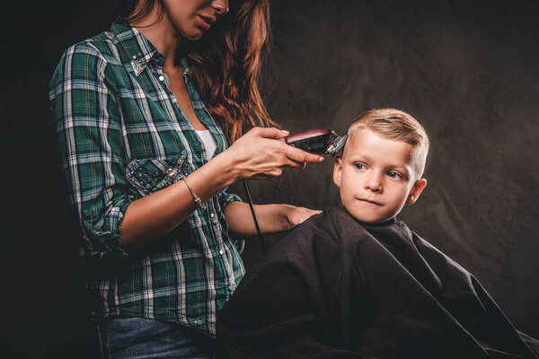 Children hairdresser with the trimmer is cutting little boy against a dark background. Cute preschooler boy getting haircut.