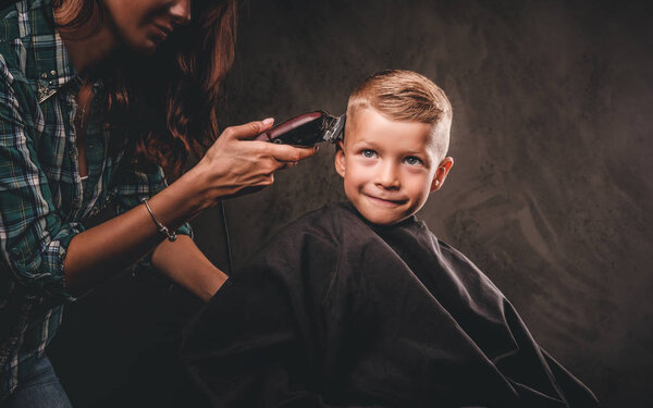 Children hairdresser with the trimmer is cutting little boy against a dark background. Contented cute preschooler boy getting haircut.