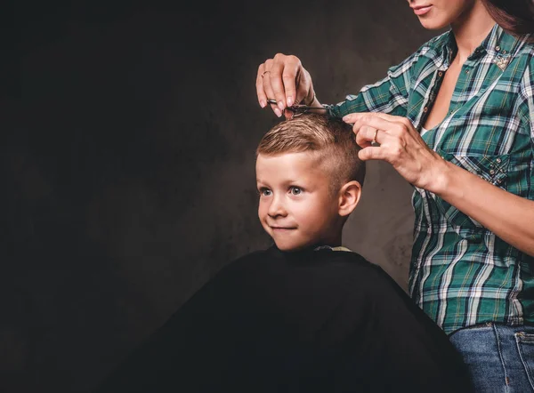 Children hairdresser with scissors is cutting little boy against a dark background. Contented cute preschooler boy getting haircut.