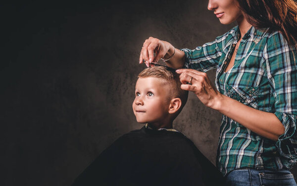 Children hairdresser with scissors is cutting little boy against a dark background. Contented cute preschooler boy getting haircut.