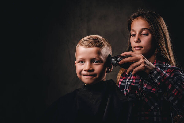The older sister cuts her little brother with a trimmer against the dark background. Cute preschooler boy getting haircut.