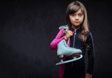 Sevimli küçük kız spor içinde giyinmiş bir omzuna buz pateni tutar. Karanlık dokulu bir arka plan üzerinde izole.