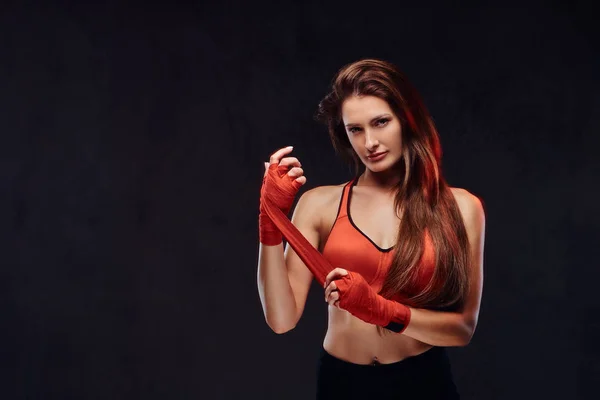 Portrait of a beautiful brunette female boxer in sports bra bandages her hands. Isolated on dark textured background.