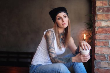 Bir hipster tattoed kız portresi paket servisi olan restoran kahve tutar ve çatı iç ile bir odada masada otururken pencereden görünüyor.
