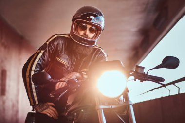 Acımasız bir sakallı motosiklet kask ve güneş gözlüğü yakın çekim portre dahil Far ile retro bir motosiklet üzerinde oturan bir siyah deri ceket giymiş.