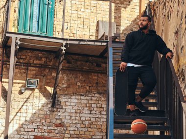 Bir siyah hoodie kılığında sakallı hippi adam koyu tenli ve spor şort bir mahallede duran bir kaykay merdiven üzerinde tutar.