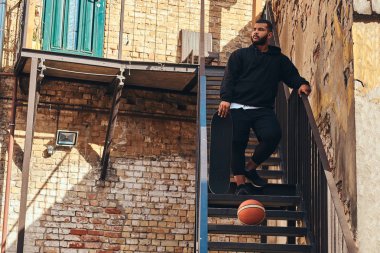 Bir siyah hoodie kılığında sakallı hippi adam koyu tenli ve spor şort bir mahallede duran bir kaykay merdiven üzerinde tutar.