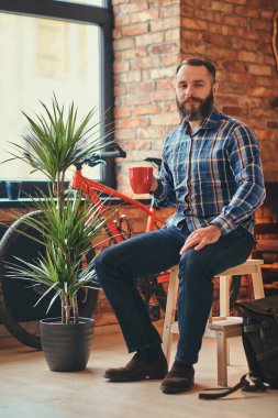 Studio loft iç ahşap bir dışkı üzerinde otururken yakışıklı sakallı hippi erkek mavi Polar gömlek ve pantolon sabah kahve tutar.