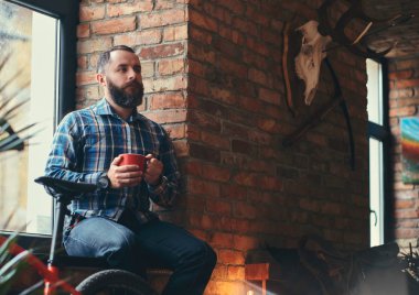 Studio loft iç pencere pervazına üzerinde otururken sabah kahve yakışıklı sakallı hippi erkek mavi Polar gömlek ve pantolon tutar.