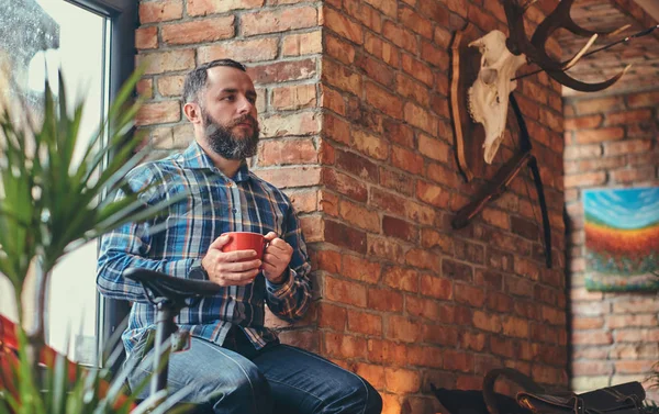 Portrait of a handsome bearded hipster male in a blue fleece shirt and jeans holds a cup of morning coffee while sitting on a window sill at a studio with loft interior.