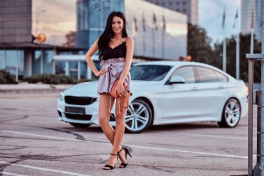 Modern giysiler giymiş baştan çıkarıcı esmer kadın çanta lüks bir araba bir gökdelen karşı yakın poz tutar.