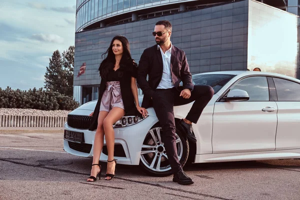 Well-dressed attractive couple leaning on a luxury car outdoors against the skyscraper. — Stock Photo, Image